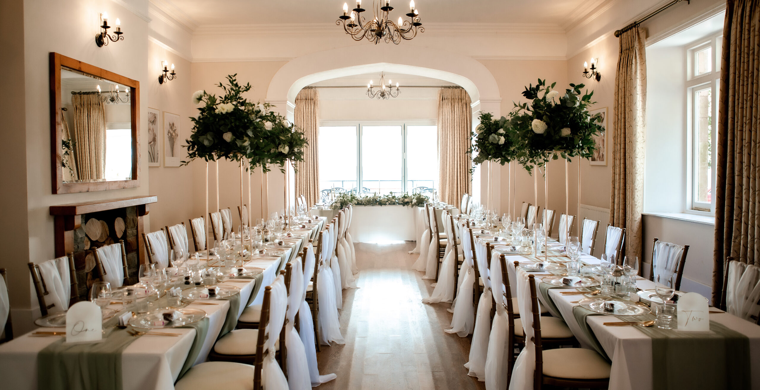 Dining room set for event; long tables with white cloths, green runners, and floral centerpieces.