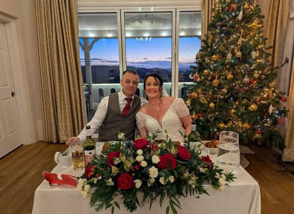 Couple at a table with floral centerpiece, Christmas tree, and twilight view through windows.