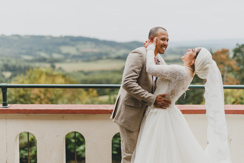 Couple laughing and embracing on terrace with countryside view, woman in white dress and headscarf