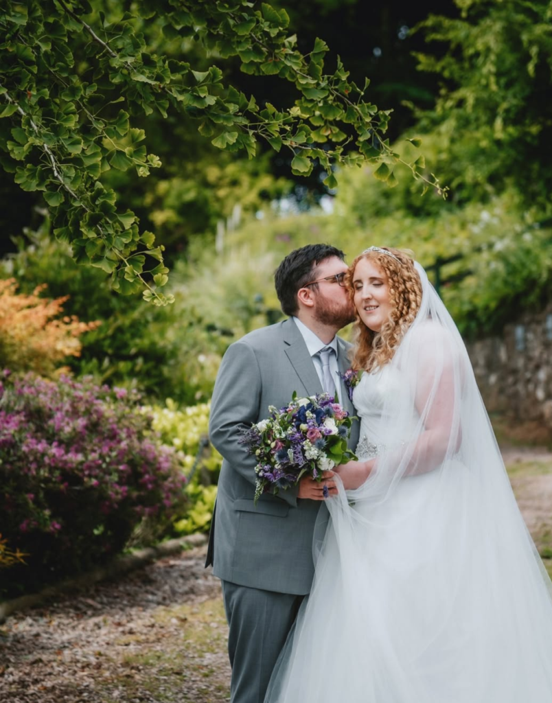 Couple in wedding attire, groom kisses bride's cheek, under leafy branches in a garden setting.